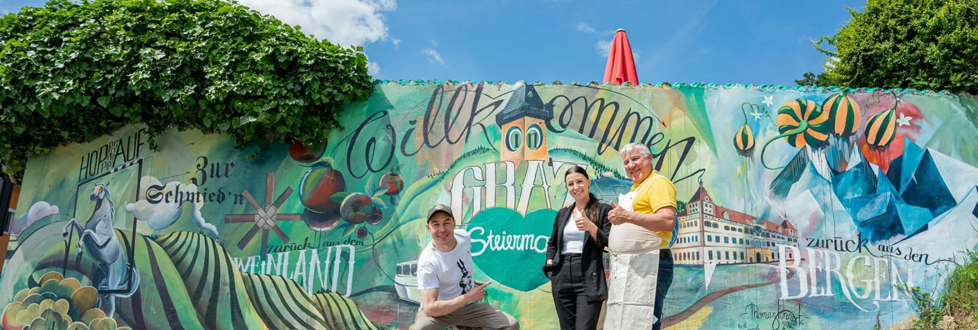 Künstler Tom Lohner und Zur Schmied´n-Gastgeber Anna-Lisa und Toni Maier mit der neuen Wandermalerei beim Gasthaus Zur Schmied´n.  | © STG | Jaden Gynes
